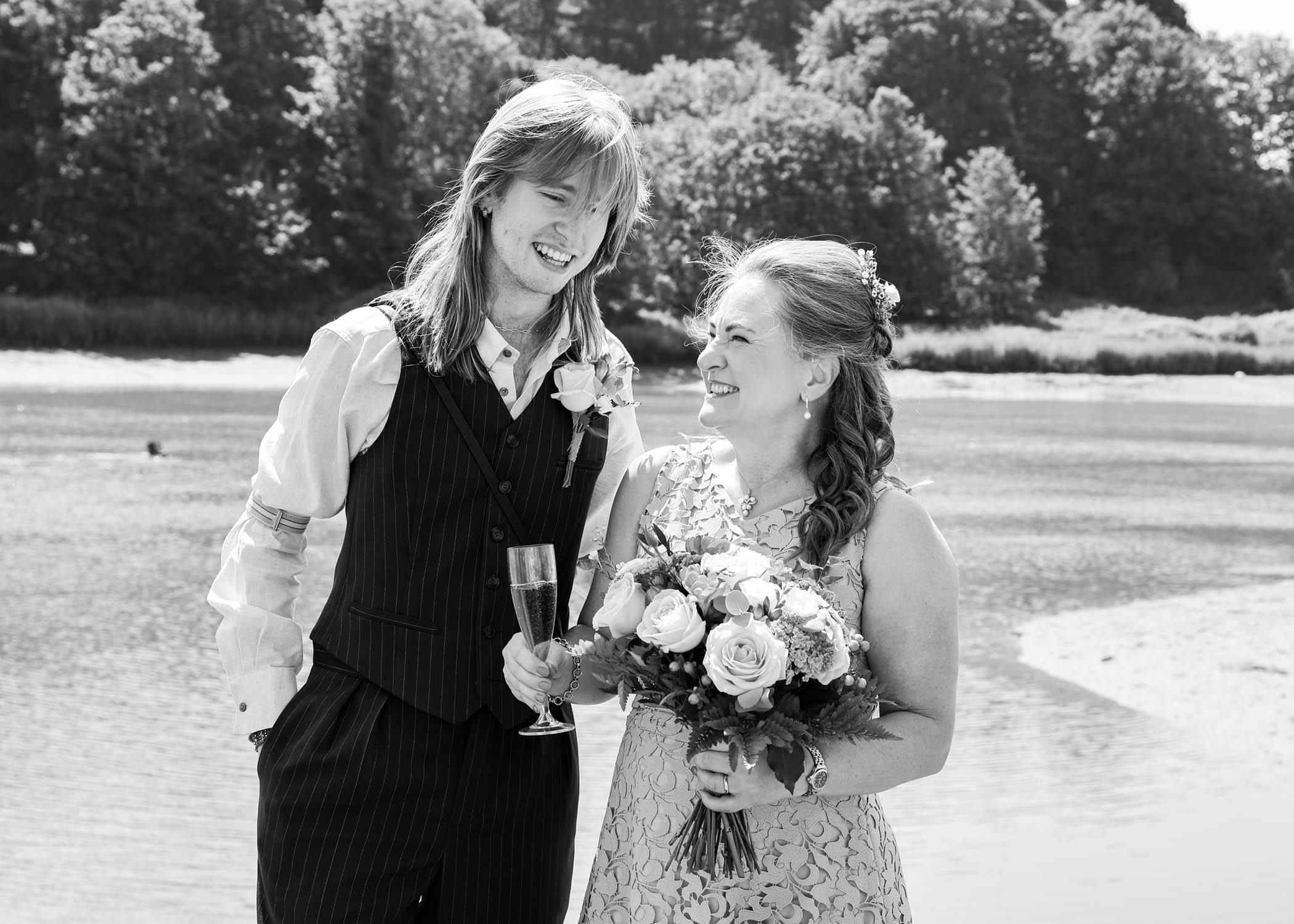 A black and white photo of a bride and her son laughing on the penisula at woodbridge marina on a wedding day in Woodbridge captured by Suffolk Wedding Photographer Hayley Denston Photography